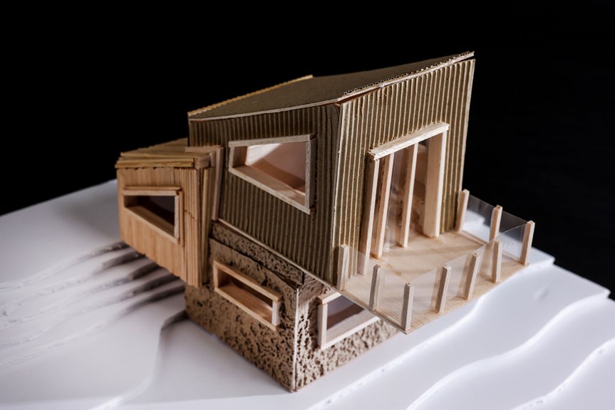 The image shows a three-dimensional architectural model of an angular cabin, made from plywood and corrugated cardboard. A balcony projects from one of its sides.