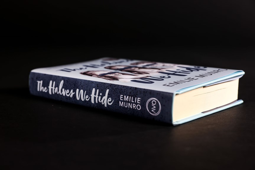 A blue book is photographed lying face up. The book’s spine reads “The Halves We Hide, Emilie Munro”. The Avo logo appears at the base of the spine.