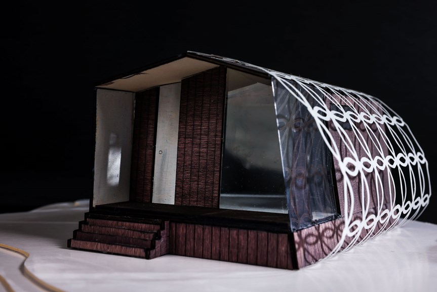 This photograph shows an architectural model of an alpine retreat. It is elevated, with four stairs leading to a doorway. The structure’s walls are made of dark wood panels, with an embellished white design feature curving out from one of its sides.