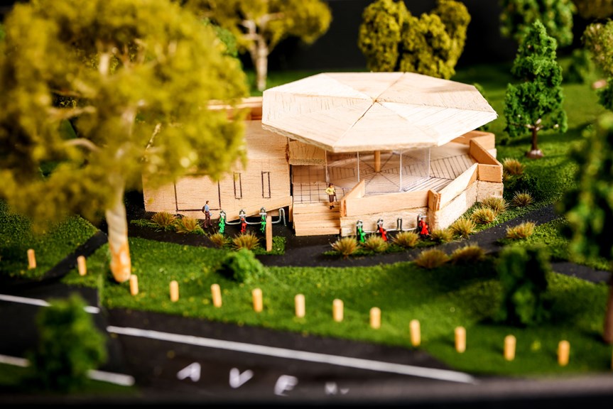 The image shows a three-dimensional architectural model of pavilion with a geometric roof. The pavilion is surrounded by gardens and has a number of bike racks at its entrance.