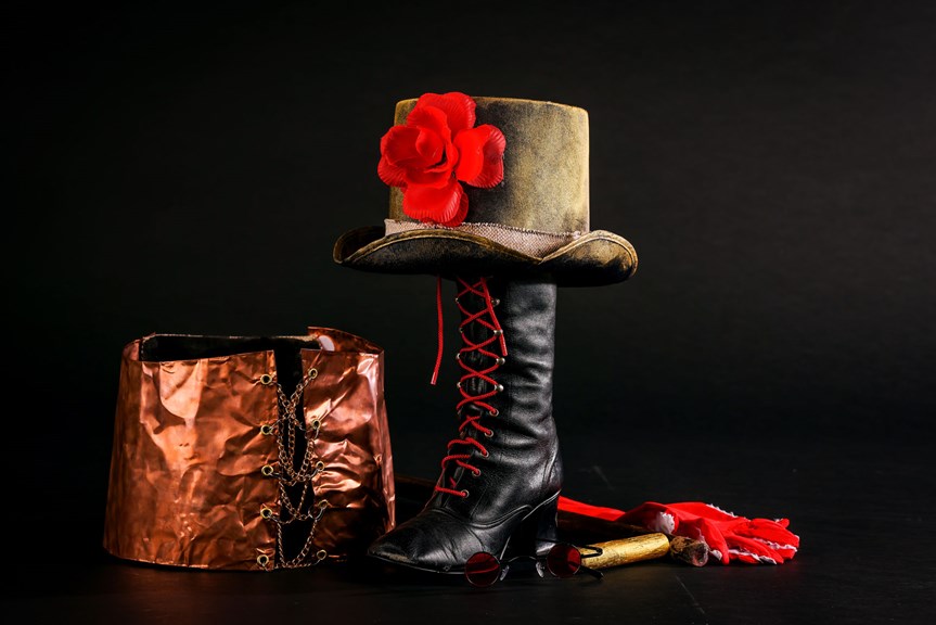 A collection of costume items: a copper corset with metal chain; a black leather boot with red laces; a brown top hat with a red flower; a pair of red and white gloves; red-tinted glasses.