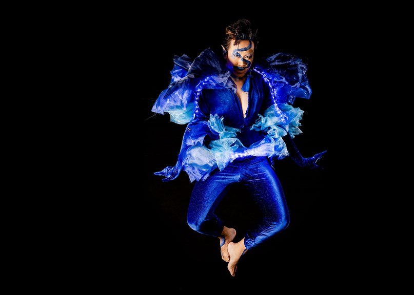 The designer is caught mid-jump modelling costume and makeup. A deep blue leotard is embellished with blue ruffles and LED lights. They have a blue waive painted across the face, pointed ears, and blue eye makeup.