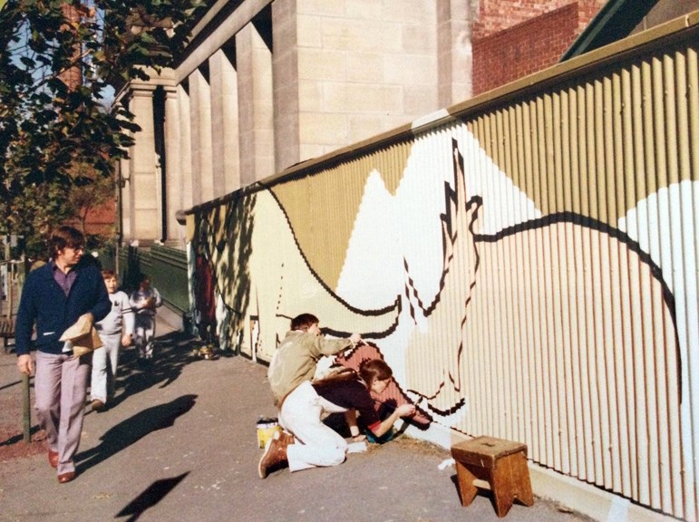 a man and a woman on their knees painting dinosaurs on a corrugated iron fence in front of a stone building, while pedestrians walk past of the footpath 