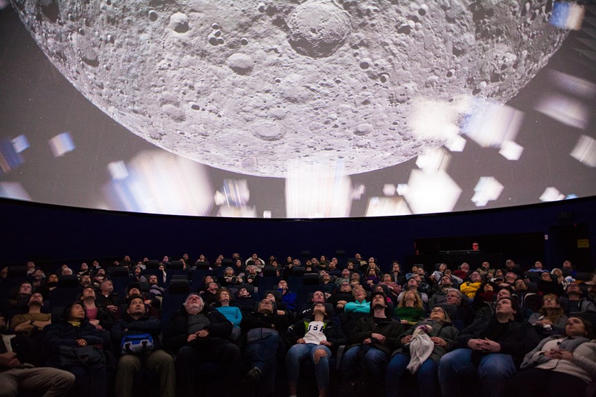 dozens of people reclining in chairs looking up at a giant projection of the moon 