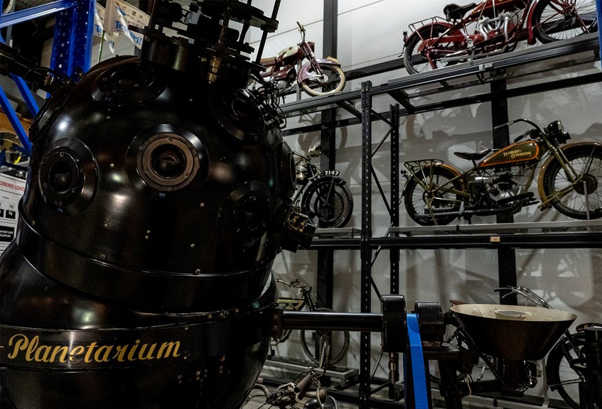 a black metal dome covered with glass lenses, with the word planetarium painted on the side in gold. A rack of motorbikes sits behind
