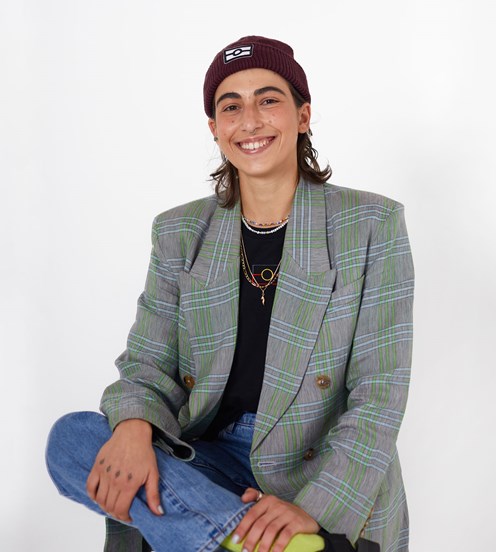 Portrait of a woman sitting on a stool. She smiling, wearing a beanie, jacket and jeans