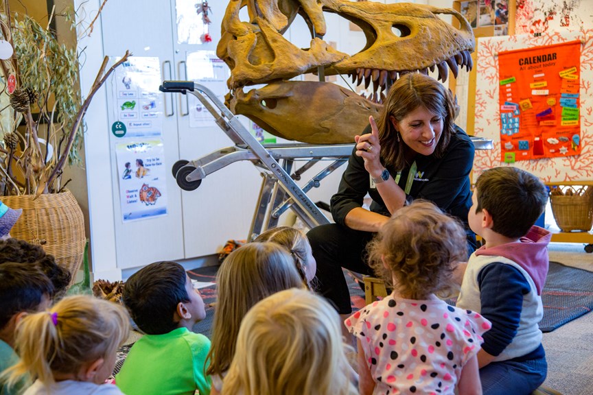 A woman sitting in front of a group of young children points at a cast of a dinosaur skull with big pointed teeth