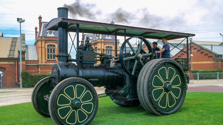 a large green-coloured engine with four wheels, in front of a big old brick building