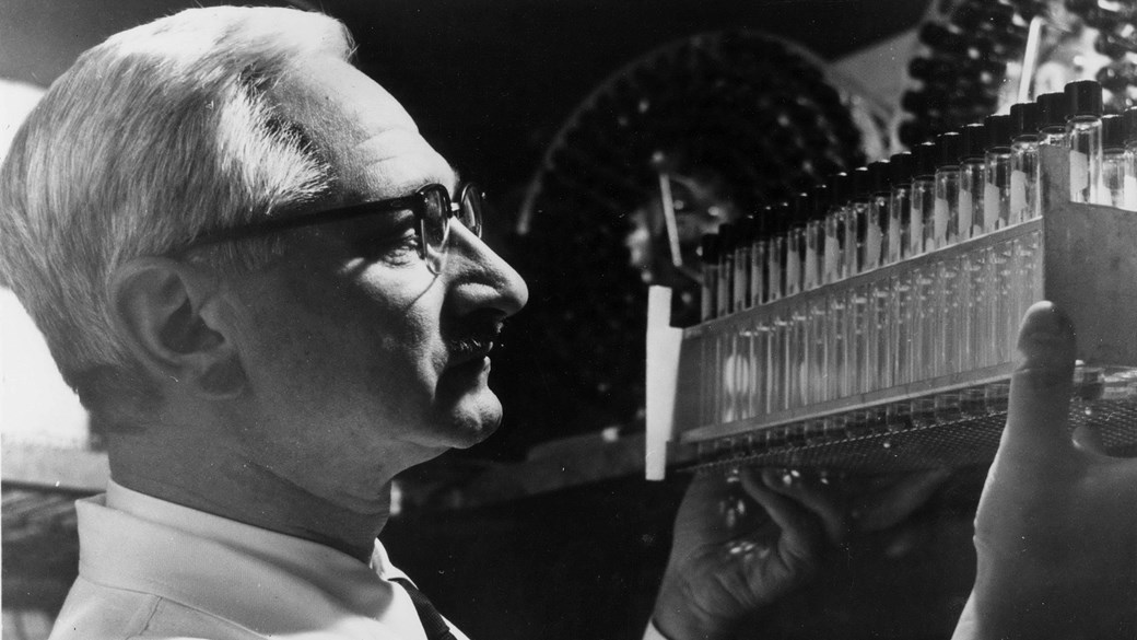 a black and white photo of a silver haired man in glasses, holding a row of glass tubes 