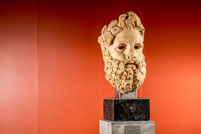 Sculpture of a bearded head perched on a plinth, against a red background