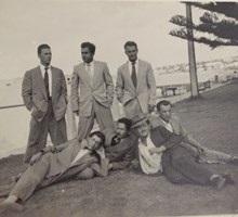 7 men in suits. Three standing, four lounging on grass.