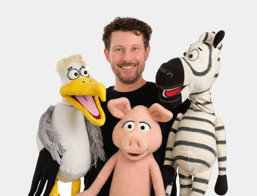  Chris Elk with a pelican, pig and zebra puppet smiling.