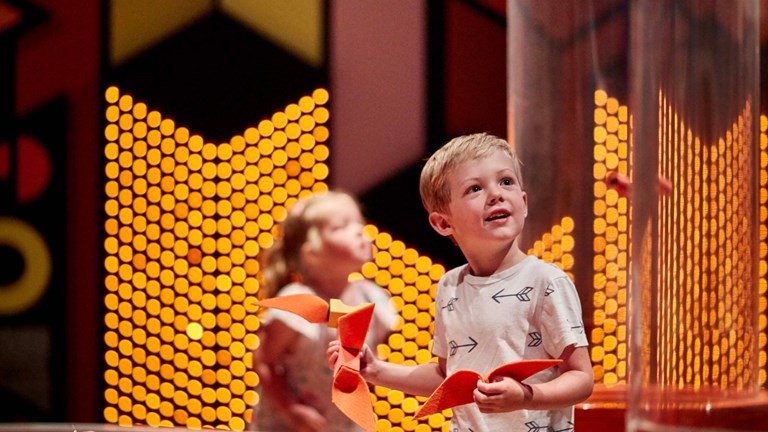 A boy holding a paper plane in a bright exhibition space. There is another child out of focus in the background.