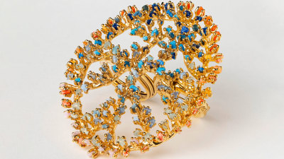 A golden ring with large flat plate on top, which is interlaced with small, bright pieces of orange, blue and green microplastic. 