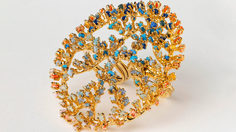 A golden ring with large flat plate on top, which is interlaced with small, bright pieces of orange, blue and green microplastic. 