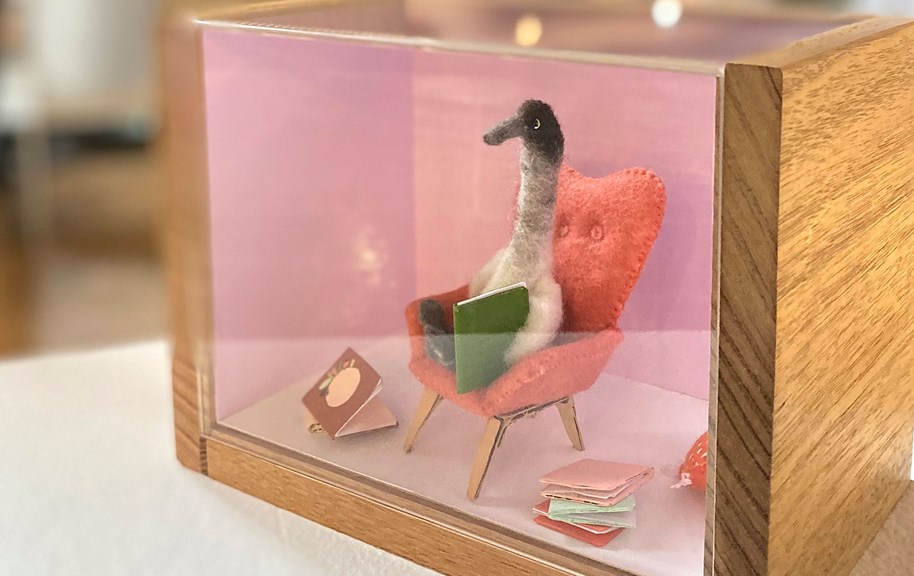 A grey goose sits in a tiny green chair reading tiny books. This scene made from felt and cardboard is a diorama behind glass in a wooden box.