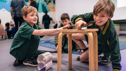 Three primary school students in uniforms are using a stool and some materials to build a marble rollercoaster during an education program at Scienceworks.  