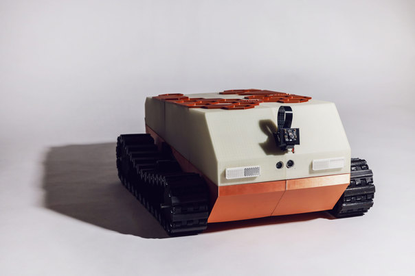3D-printed tracked vehicle in cream and terracotta colours featuring a sloped top with solar panels, dual tank treads, and visible electronic components on the front.