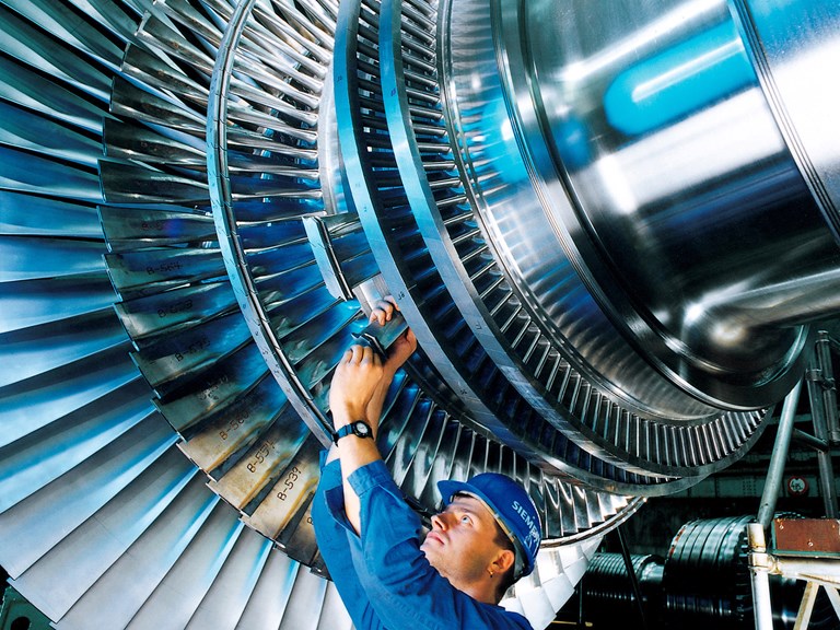 a man wearing blue overalls and a hard hat reaching up to place a blade on a large metal turbine
