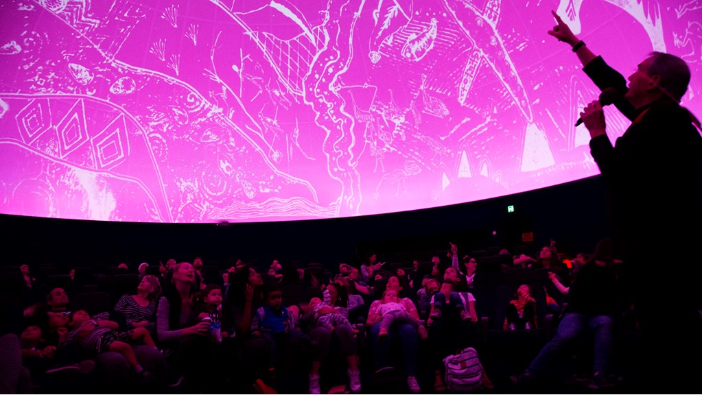 Audience members at the Planetarium are seat and looking up at a bright pink dome while a presenter explains the constellations depicted on the Boorong possum skin cloak