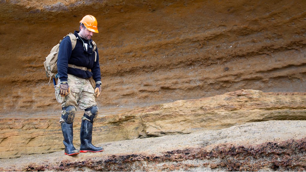 Man wearing a backpack and protective clothing standing near a beach side cliff