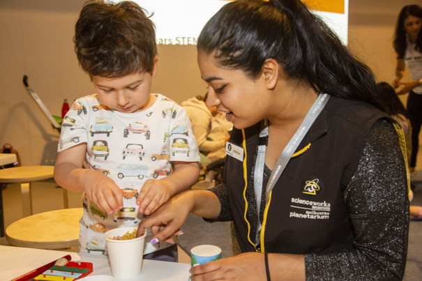 A woman with black hair in a ponytail sitting at a work table with a young boy with brown hair looking into a white cup full of things to create sound