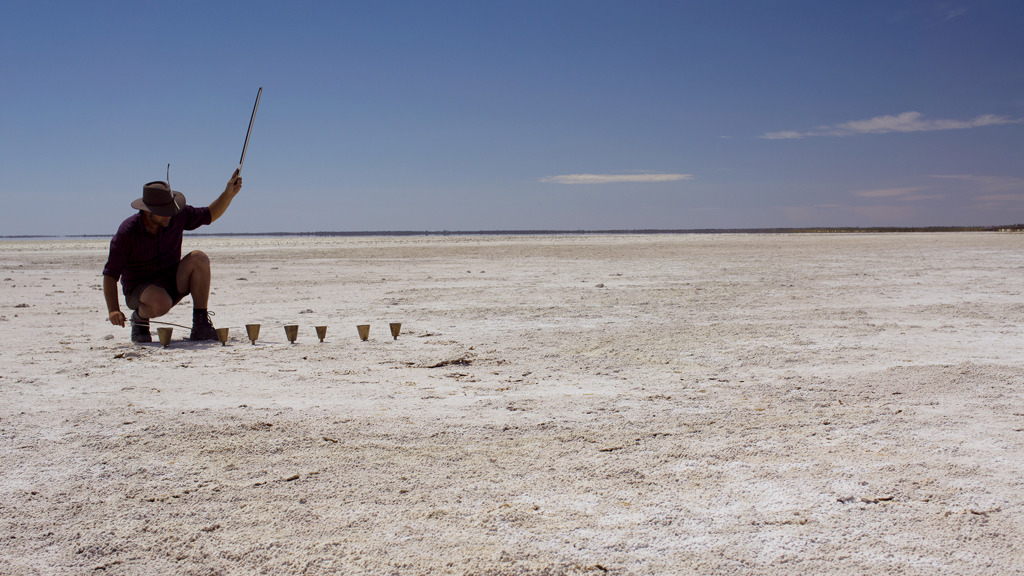 A person is crouched over a row of small bells on a salt lake. He holds a stick aloft.
