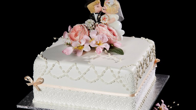 A white cake with flowers and small figurines of a suited man and woman wearing a white dress