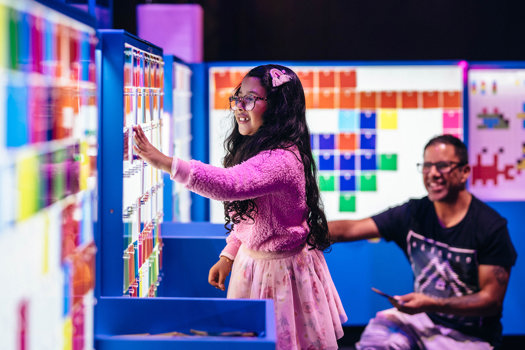 A child adds a transparent coloured square to a wall as a parent looks on.