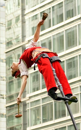 Acrobat on a stilt, bent over backwards, holding in one hand a mallet and in the other, a bell