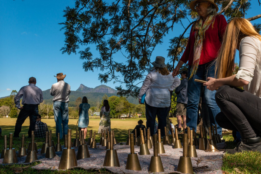Many handbells are standing on a rug on the ground in a field on a sunny day in the countryside. Several people stand or are seated, mostly with their backs turned to the camera.