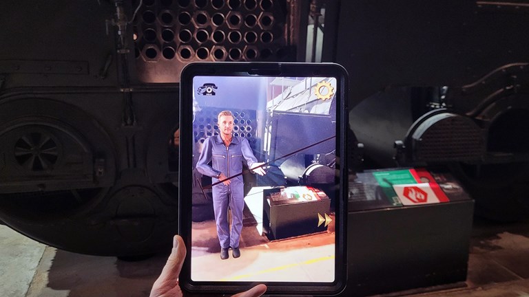 A hand holds an iPad showing an Augmented reality character in front of two large black boilers 
