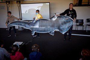 Museums Victoria personnel delivering a presentation during the Museums Victoria outreach program, with two children helping to hold up a large printed Koolasuchus cleelandi graphic