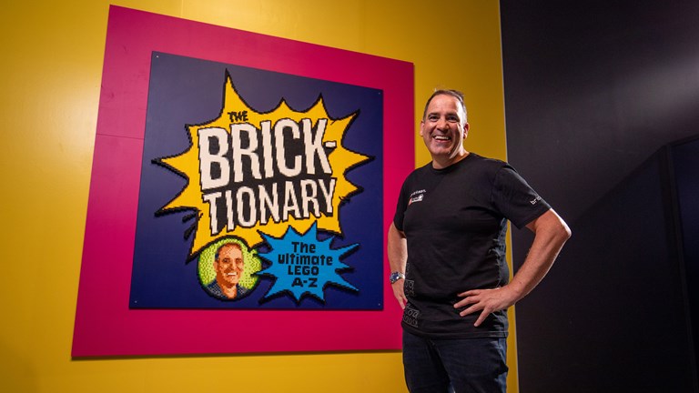 A man dressed in black with hands on hips smiles alongside a framed poster that reads "The Bricktionary: The Ultimate Lego A-Z"