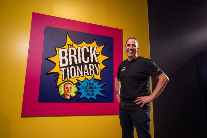 A man dressed in black with hands on hips smiles alongside a framed poster that reads "The Bricktionary: The Ultimate Lego A-Z"