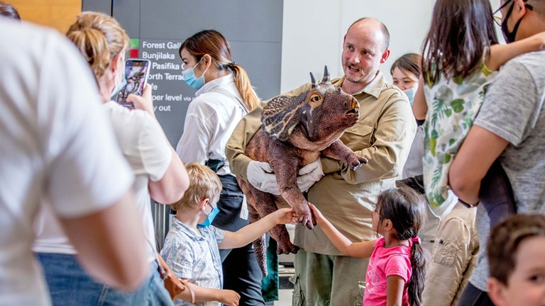 Crowd gather around a man holding a baby Triceratops puppet