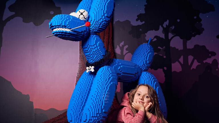 A blue Lego dog with a girl lying beneath it, head in hands. There is a woodlands backdrop.