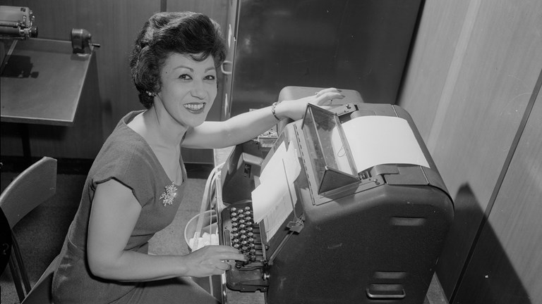 A female office worker operating a paper tape printer unit at a desk in an office