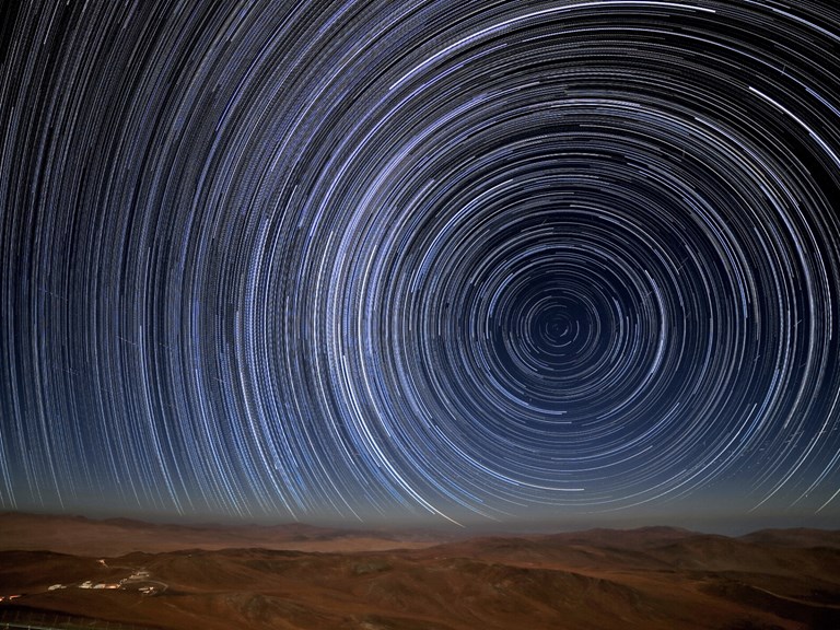 A view from the desert landscape around ESO's Paranal Observatory in Chile reveals stars' paths — star trails — as they appear to journey through the night sky. Caused by the Earth's rotation relative to a static sky, the stars appear to travel around one fixed point — Earth's Celestial South Pole — which is just visible above the rust-tinted horizon.