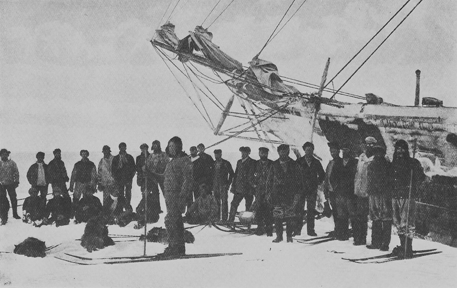 a black and white photo of a group of warmly dressed men standing on ice in front of a ship. Several dogs lie at their feet