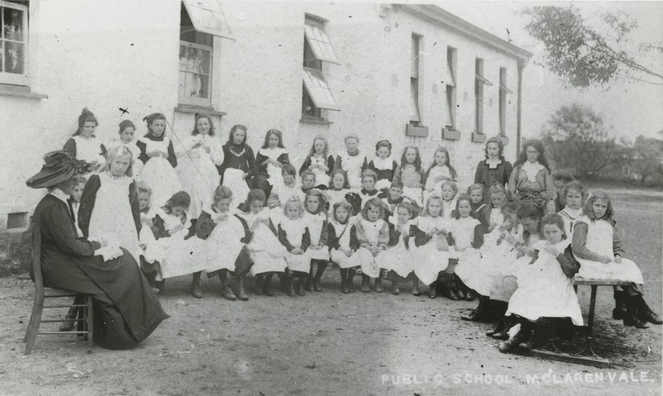 Many uniformed young girls taking an outdoors sewing class, overseen by a teacher