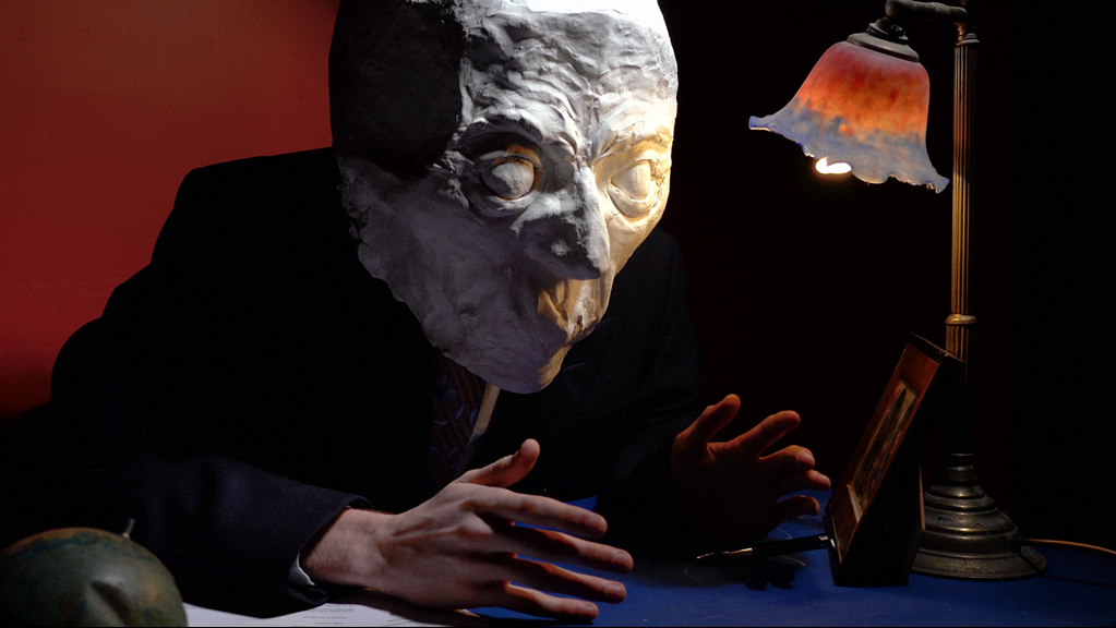 A person sat down at a desk wearing a suit and an oversized paper mache head.