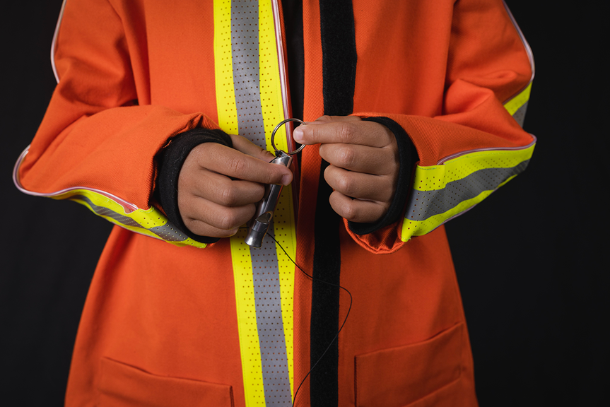 A close-up photo shows a person wearing a long-sleeved orange jacket with reflective stripes along the sleeves and front zip. Their hands hold a silver whistle in front of them. 