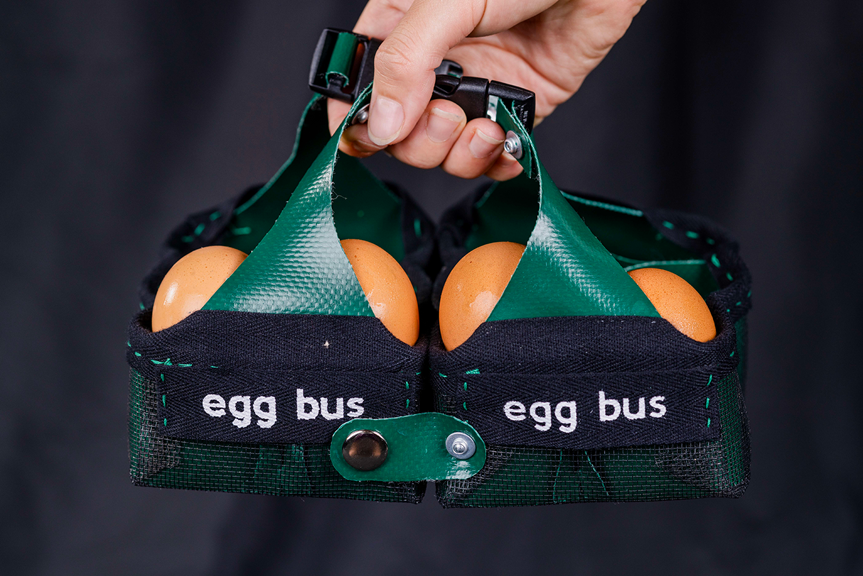 A hand holds a small green and black bag, which contains eggs. The side of the bag has white text reading ‘egg bus’.