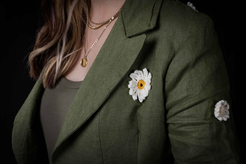 A close-up photo of a person wearing a golden necklace and dark green blazer, which has two white crocheted flowers attached to the front and sleeve.