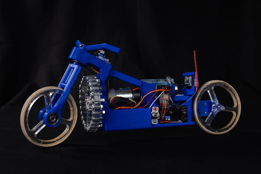 A landscape photograph showing a blue motorbike model made with various components. Coloured wires and an on/off switch are visible on its side.