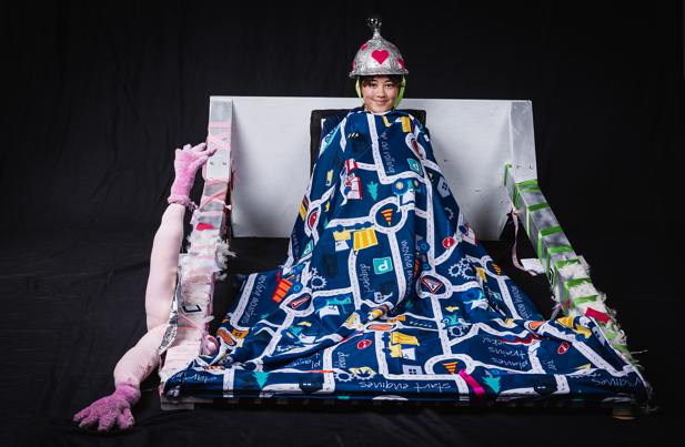 A person who sits upright against a silver frame, covered by a patterned blanket. They wear a tin foil helmet on their head that has a light bulb attached to it.