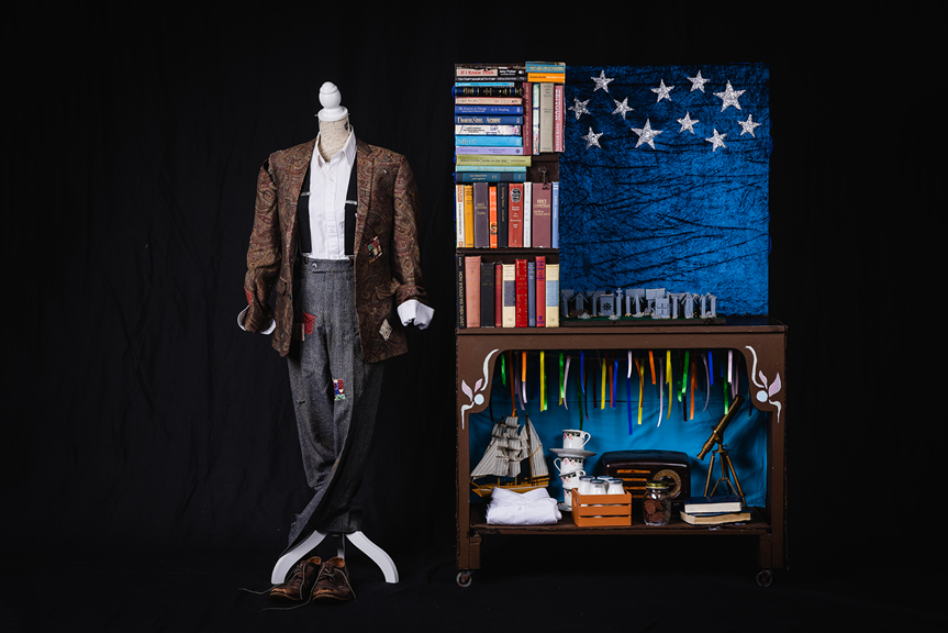 A mannequin dressed in a paisley patterned suit jacket, white shirt, braces and grey trousers. Next to the mannequin is a desk with numerous books and kick knacks placed on top of it.