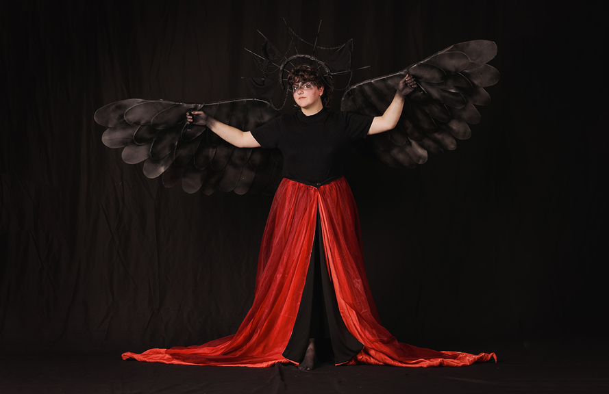 A person wears a long red skirt which is draped on the floor, with large black wings and a halo attached to their arms and head.