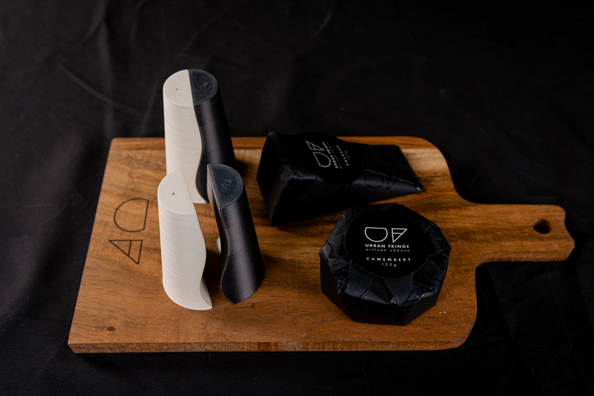 Two blocks of cheese and salt and pepper shakers sat on a wooden charcuterie board. The cheese is wrapped in black Urban Fringe packaging.